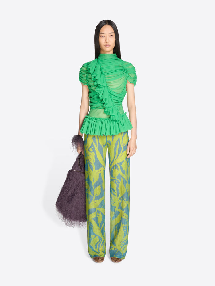 Model wearing a green ruffled top and patterned pants with a dark bag on a white background - Dries Van Noten