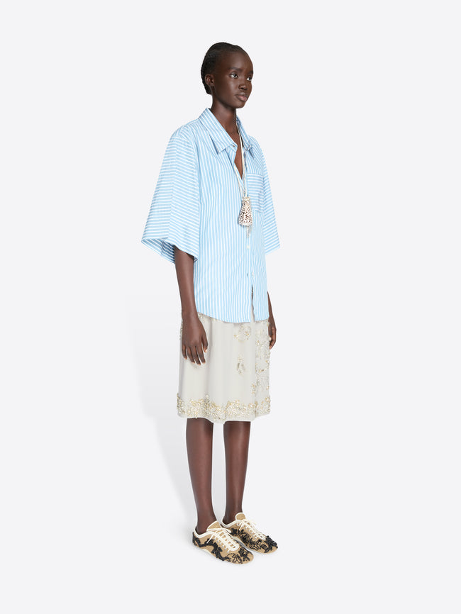 Woman wearing a light blue striped shirt and beige skirt on a white background - Dries Van Noten