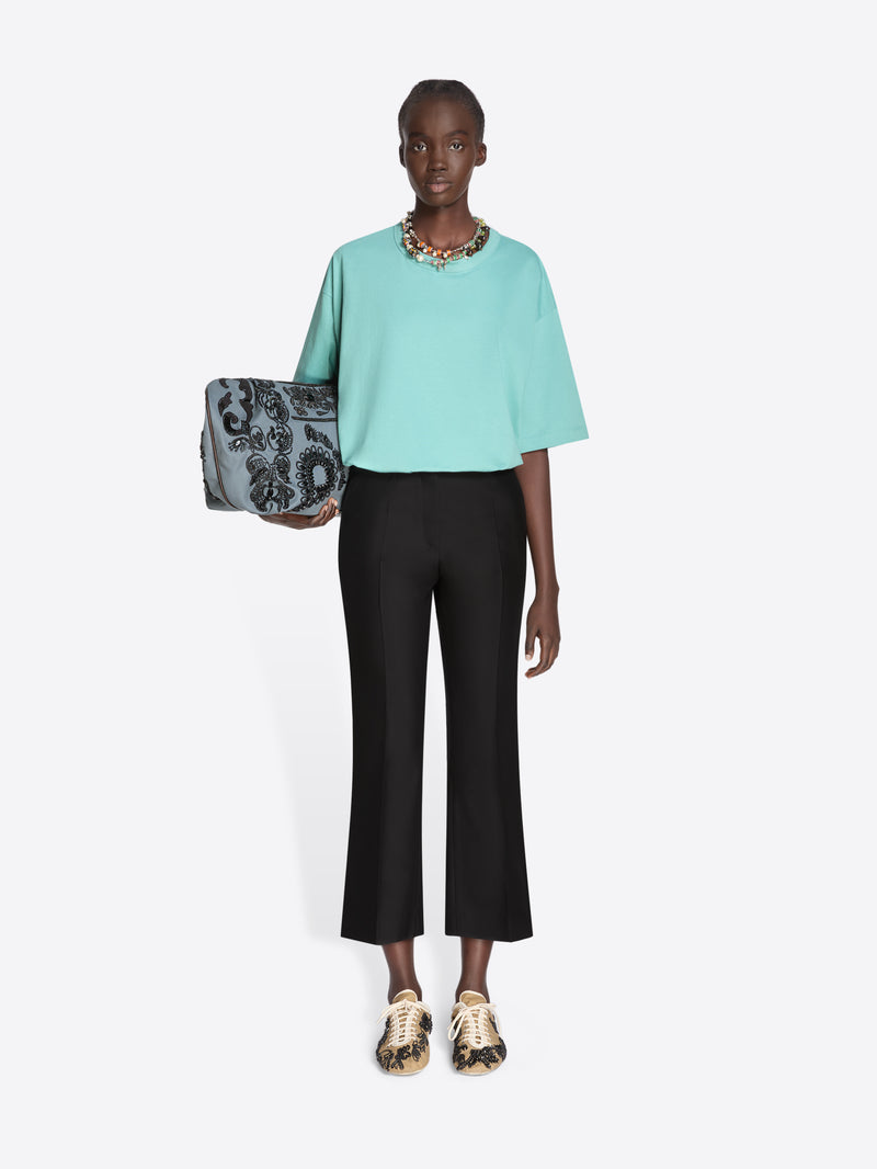 Woman wearing a light blue top and black pants, holding a patterned handbag on a white background - Dries Van Noten