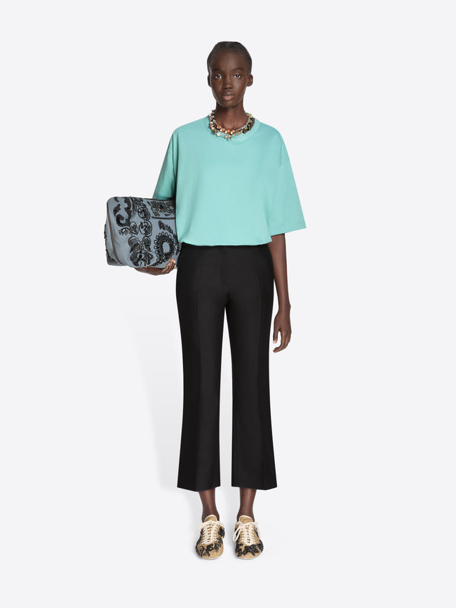 Woman wearing a light blue top and black pants, holding a patterned handbag on a white background - Dries Van Noten