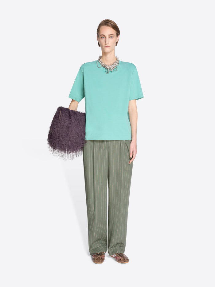 Woman wearing a light blue top and green pants holding a dark purple bag on a white background - Dries Van Noten