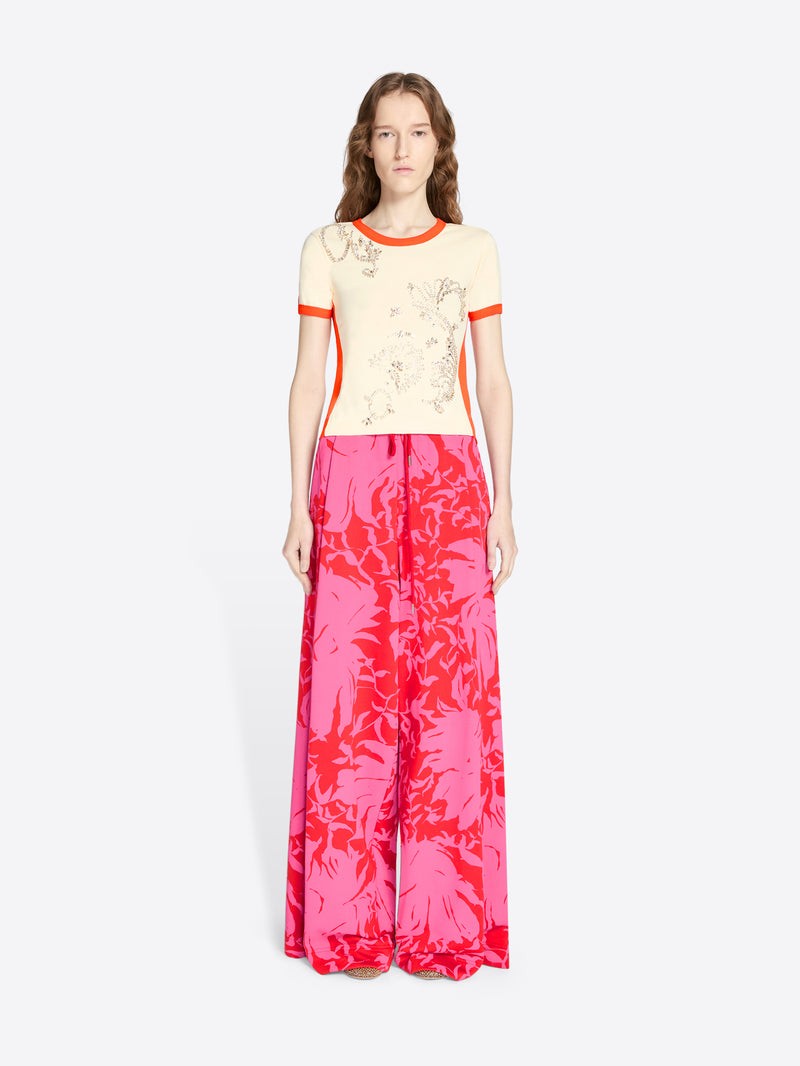 Woman wearing a cream top with orange trim and pink floral pants on a white background - Dries Van Noten