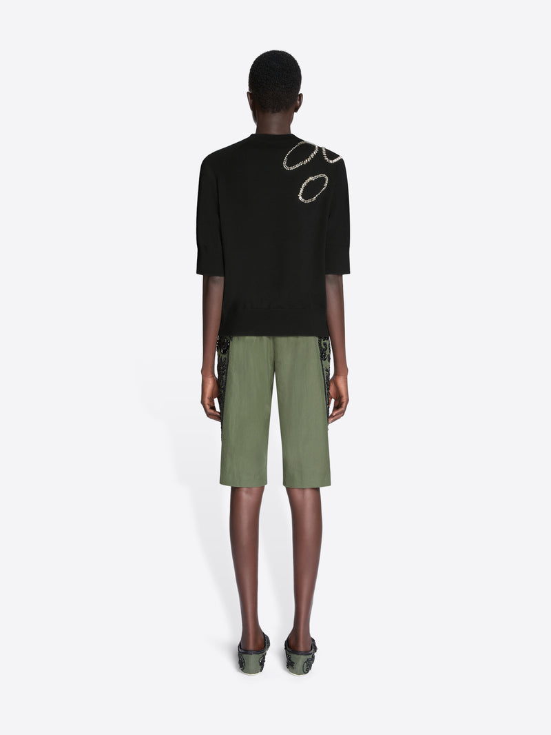 Person wearing a black t-shirt with a logo and green shorts on a white background - Dries Van Noten