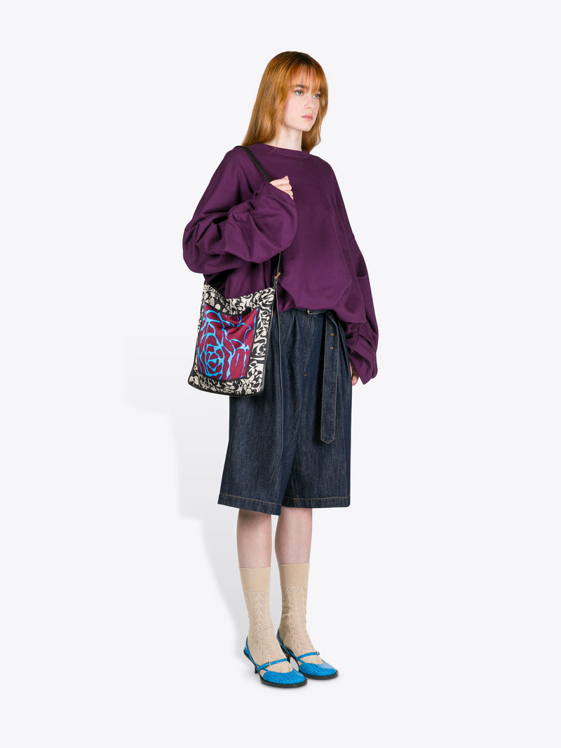 Person wearing a purple sweater and denim skirt holding a patterned bag on a white background - Dries Van Noten