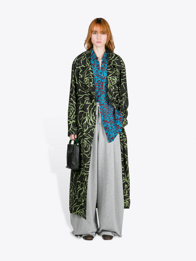 Person wearing a long black coat with green patterns, a blue patterned shirt, and gray pants on a white background. - Dries Van Noten