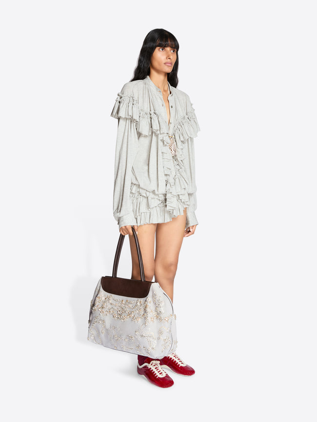Woman holding a large tote bag wearing a light-colored outfit with ruffled details on a white background - Dries Van Noten