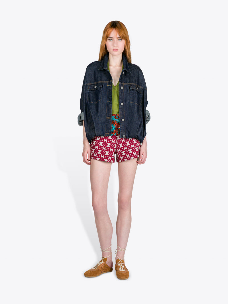 Person wearing a denim jacket, green top, patterned shorts, and brown shoes on a white background