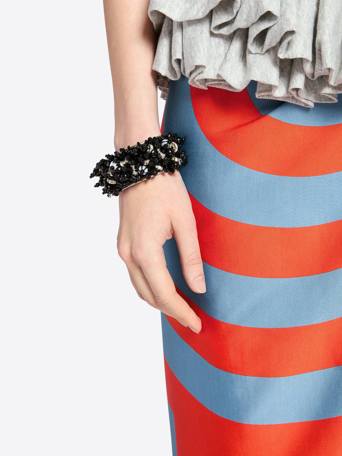 Person wearing a colorful dress with red and blue stripes, holding a black beaded bracelet. - Dries Van Noten