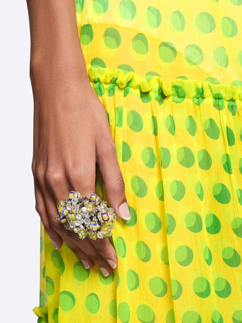 Hand wearing a decorative ring on a yellow and green polka dot fabric background - Dries Van Noten