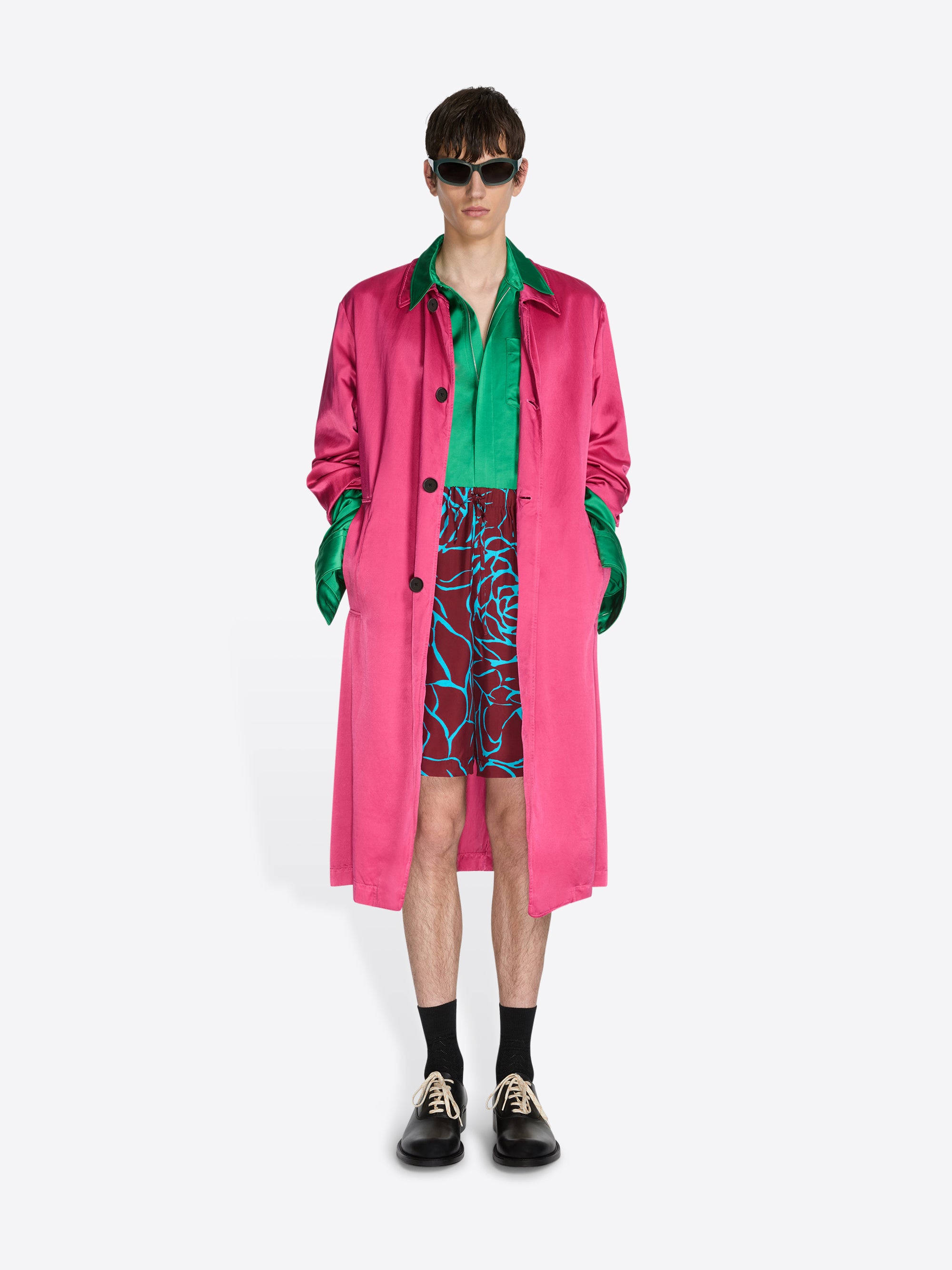 Person wearing a bright pink coat over a green shirt with a colorful patterned shorts on a white background - Dries Van Noten