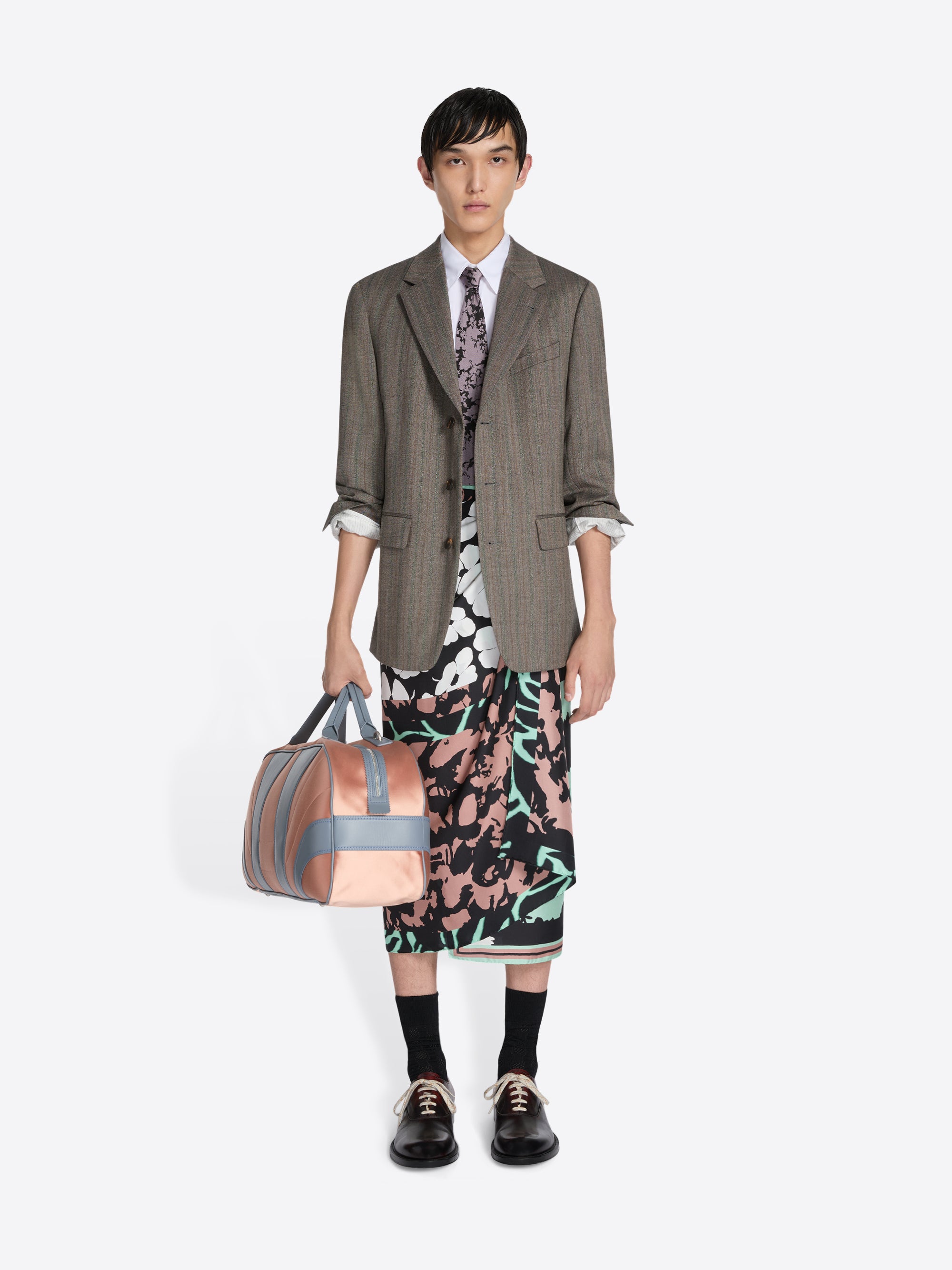 Person wearing a patterned outfit with a jacket and skirt, holding a bag on a white background