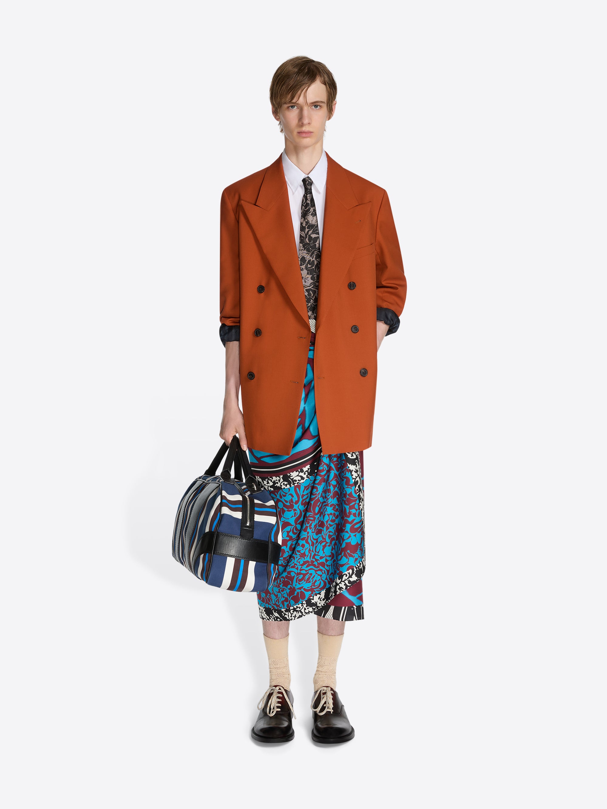 Person wearing a brown coat and colorful outfit holding a bag on a white background - Dries Van Noten
