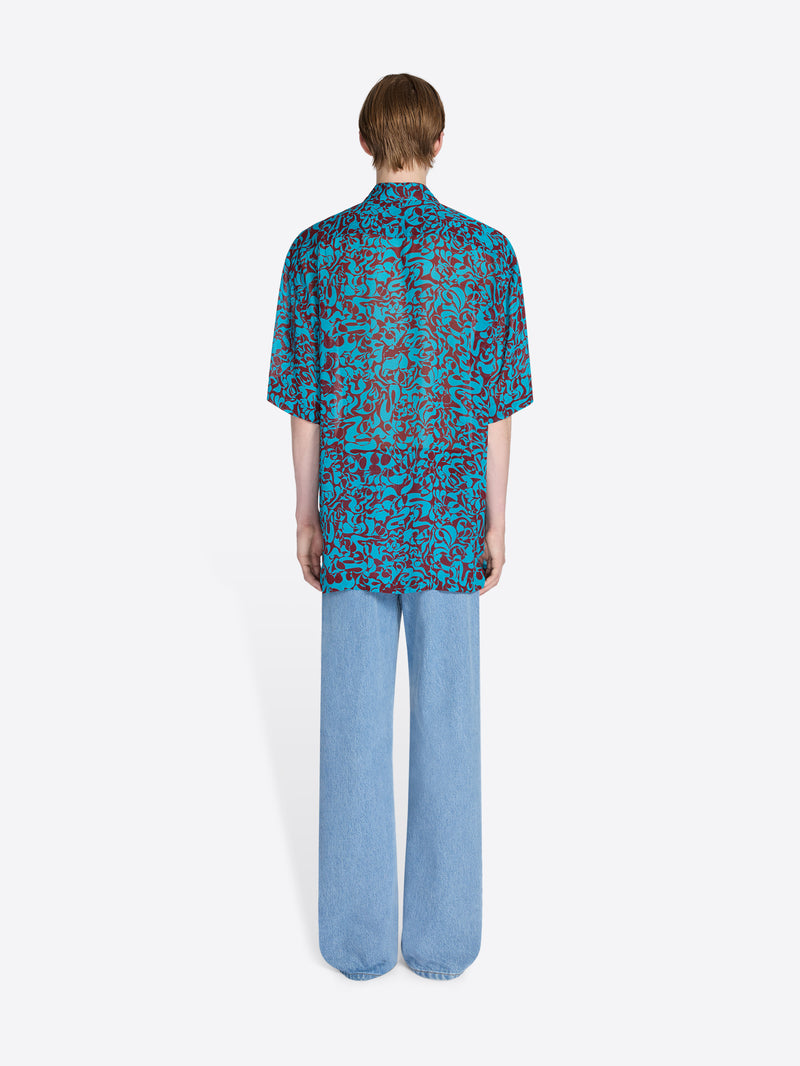 Person wearing a blue patterned shirt and light blue pants on a white background - Dries Van Noten