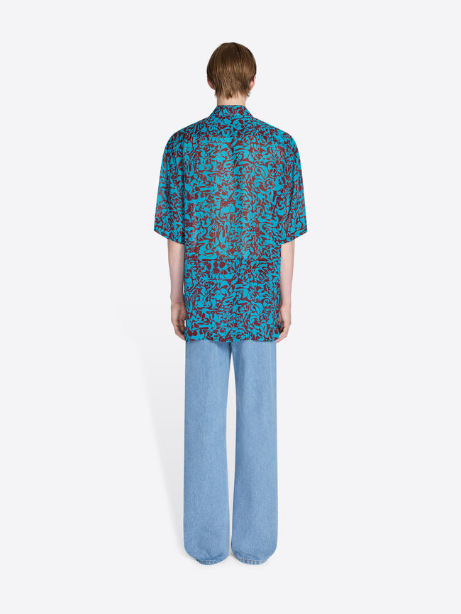 Person wearing a blue patterned shirt and light blue pants on a white background - Dries Van Noten