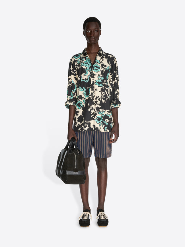 Person wearing a floral shirt and striped shorts, holding a black bag on a white background - Dries Van Noten
