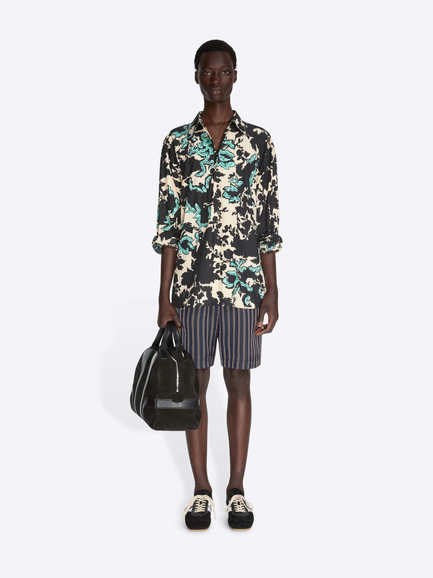 Person wearing a floral shirt and striped shorts, holding a black bag on a white background - Dries Van Noten