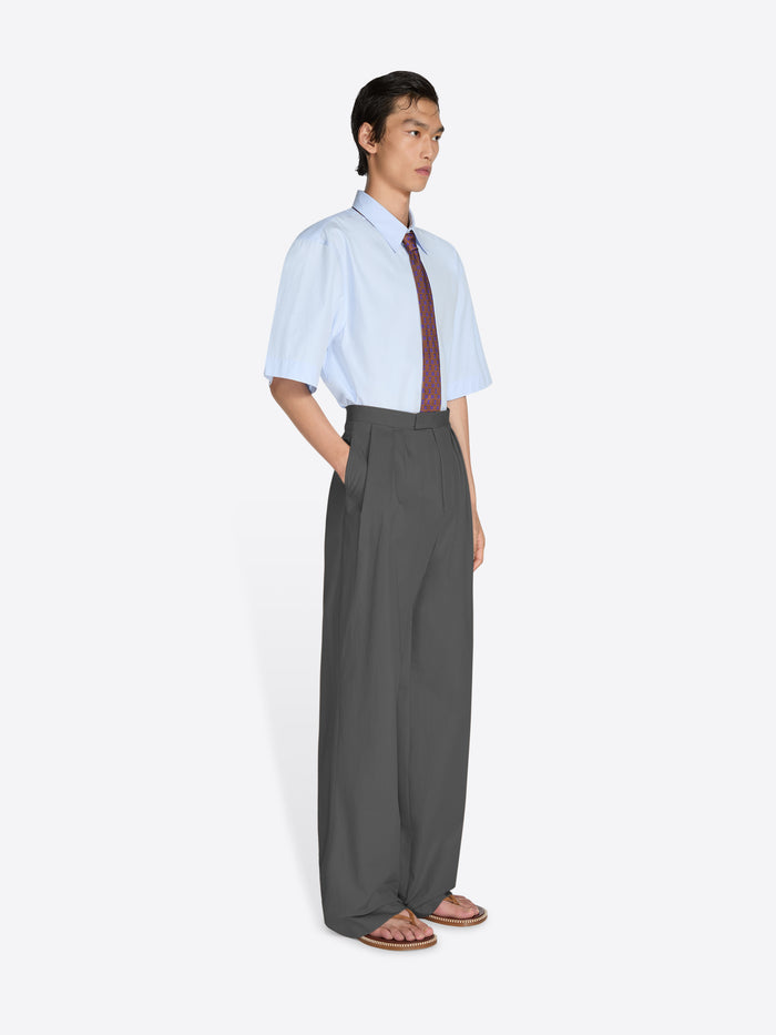 Person wearing a light blue shirt, dark tie, and gray wide-leg pants on a white background - Dries Van Noten