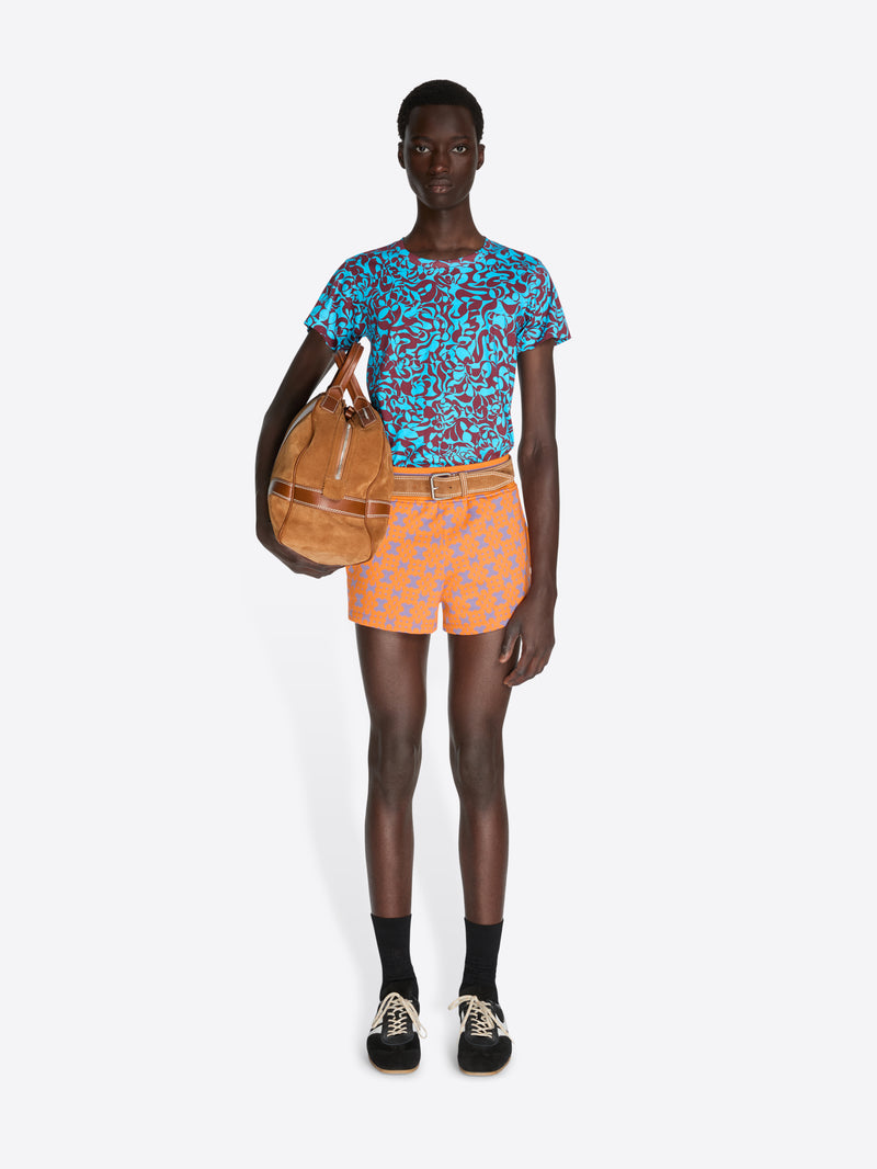 Person wearing a patterned blue shirt and orange shorts, holding a brown bag on a white background