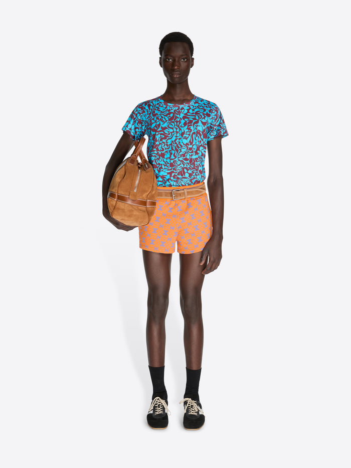 Person wearing a patterned blue shirt and orange shorts, holding a brown bag on a white background