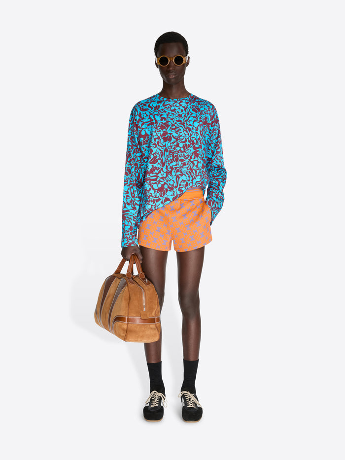 Person wearing a colorful outfit with a blue patterned top and orange shorts, holding a brown bag on a white background.