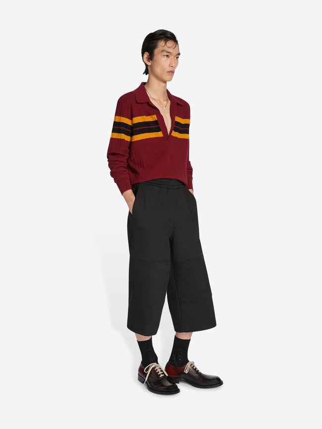 Person wearing a burgundy sweater with yellow stripes and black pants on a white background