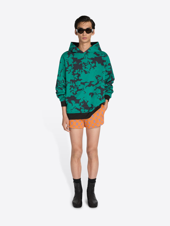 Person wearing a green camouflage hoodie and orange shorts on a white background - Dries Van Noten