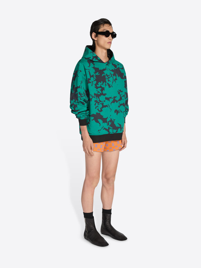 Person wearing a green camouflage hoodie and patterned shorts on a white background - Dries Van Noten
