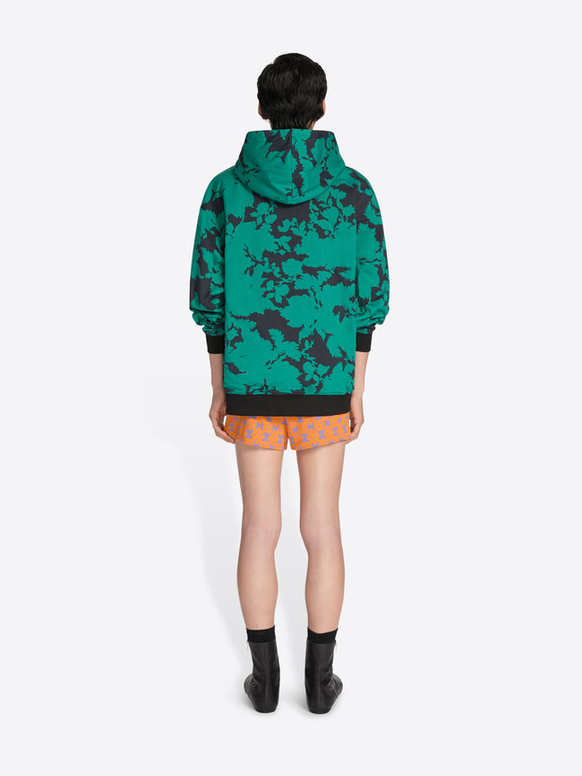Person wearing a green camouflage hoodie and orange shorts on a white background - Dries Van Noten
