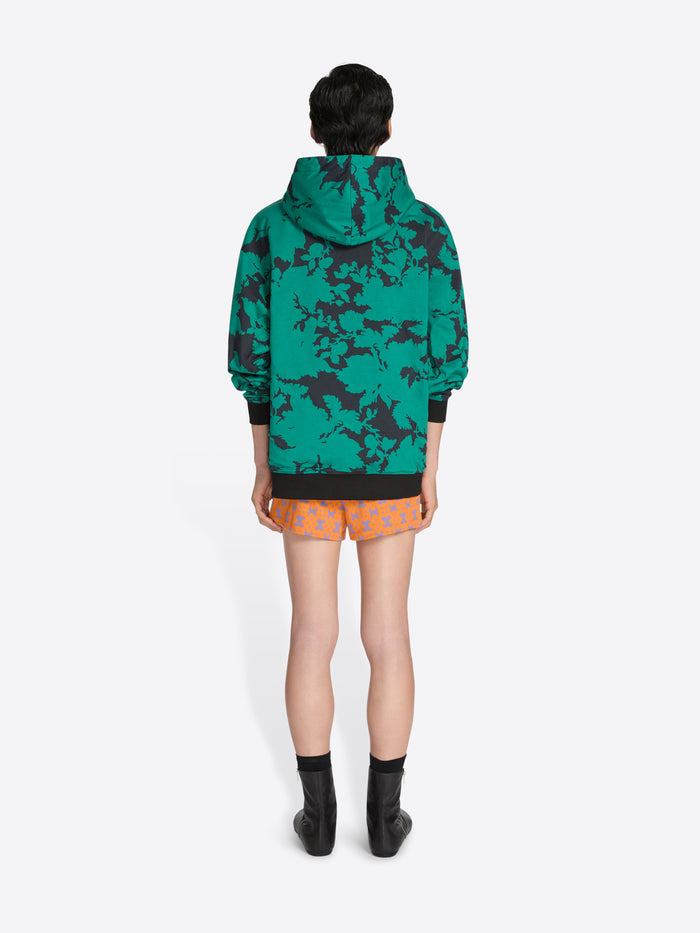 Person wearing a green camouflage hoodie and orange shorts on a white background - Dries Van Noten