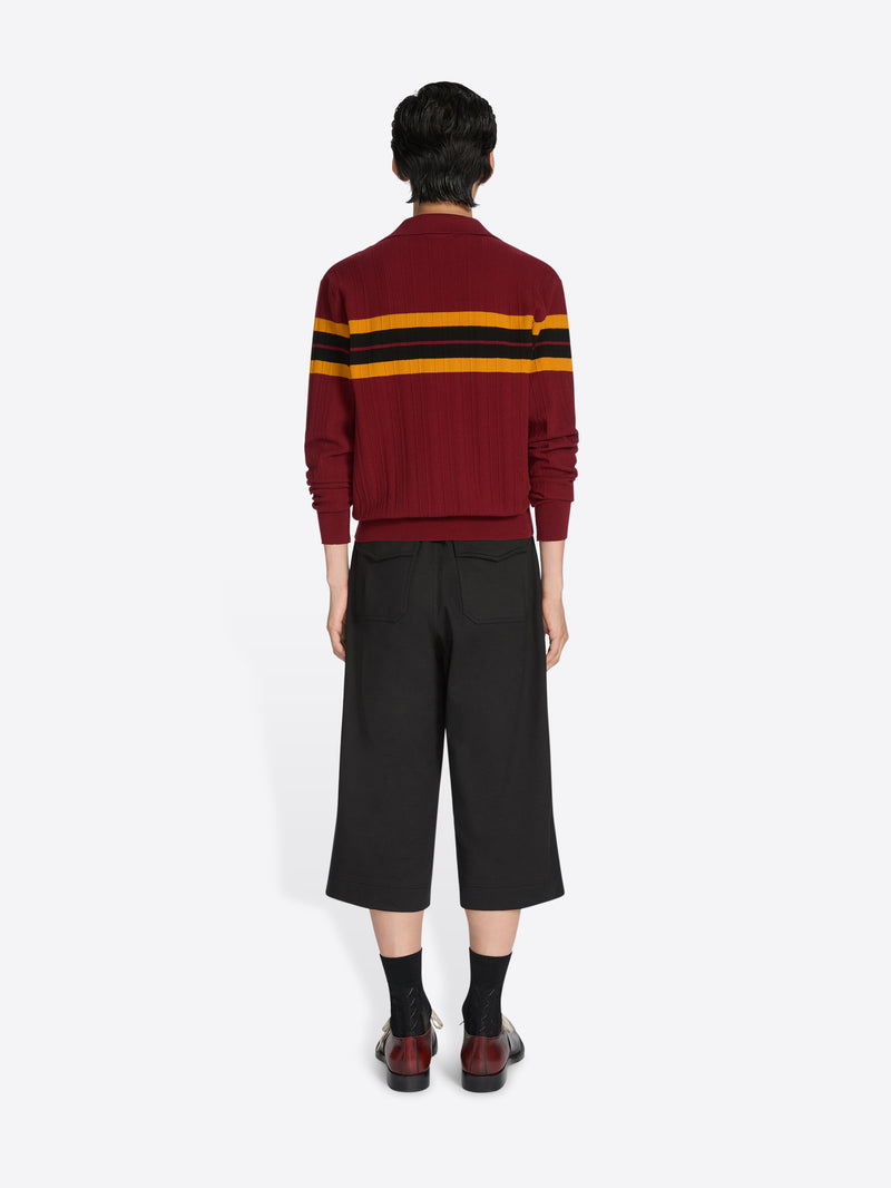 Person wearing a red sweater with yellow and black stripes and black pants on a white background - Dries Van Noten