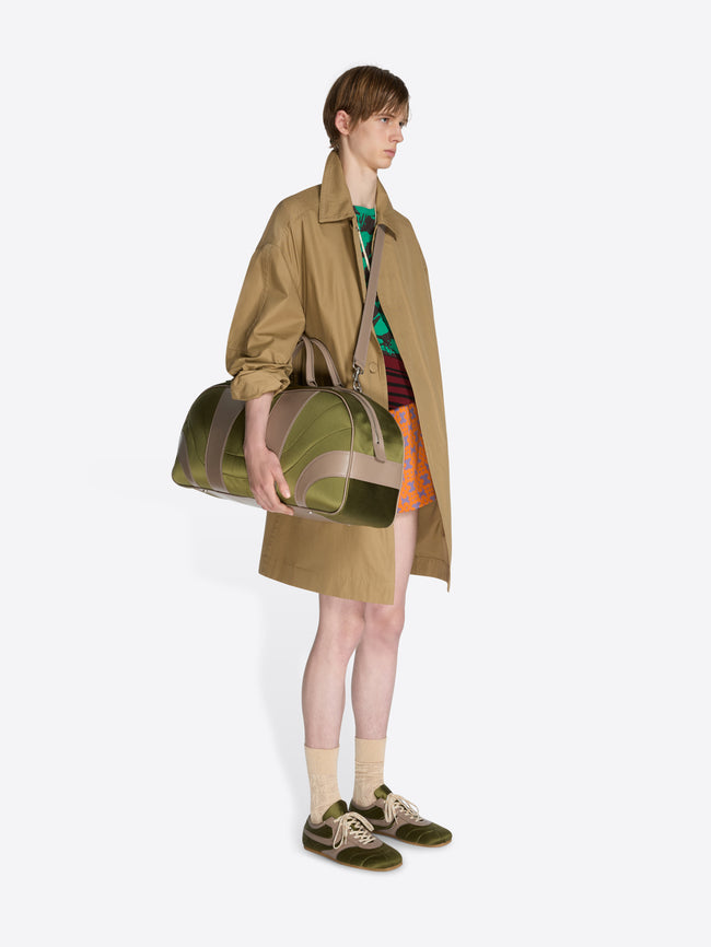 Person holding a green and brown duffel bag wearing a beige trench coat on a white background - Dries Van Noten