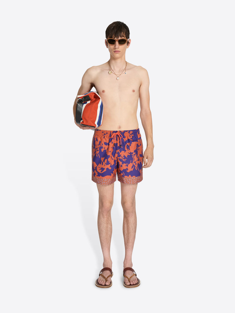 Man wearing colorful swim shorts holding a bag on a white background
