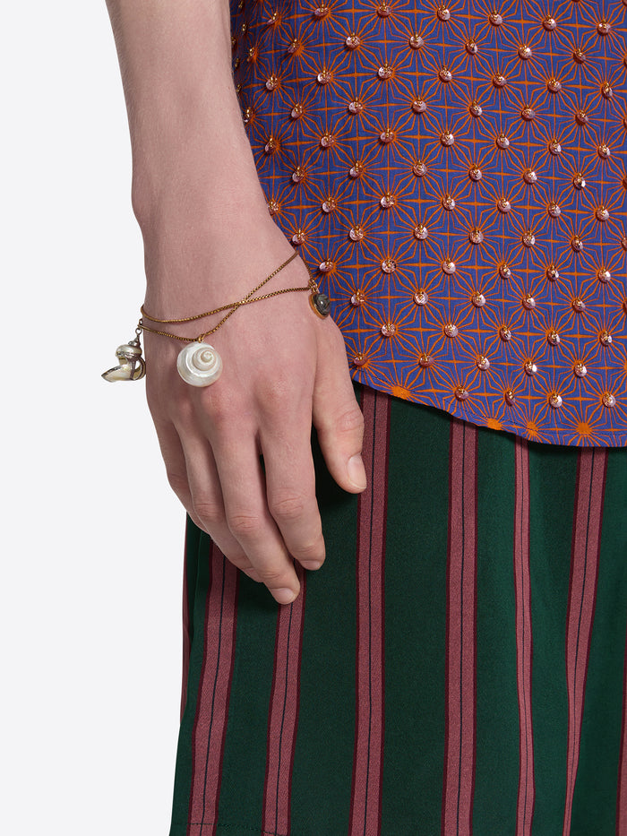 Close-up of a hand wearing a bracelet with a shell charm, against a patterned fabric background.