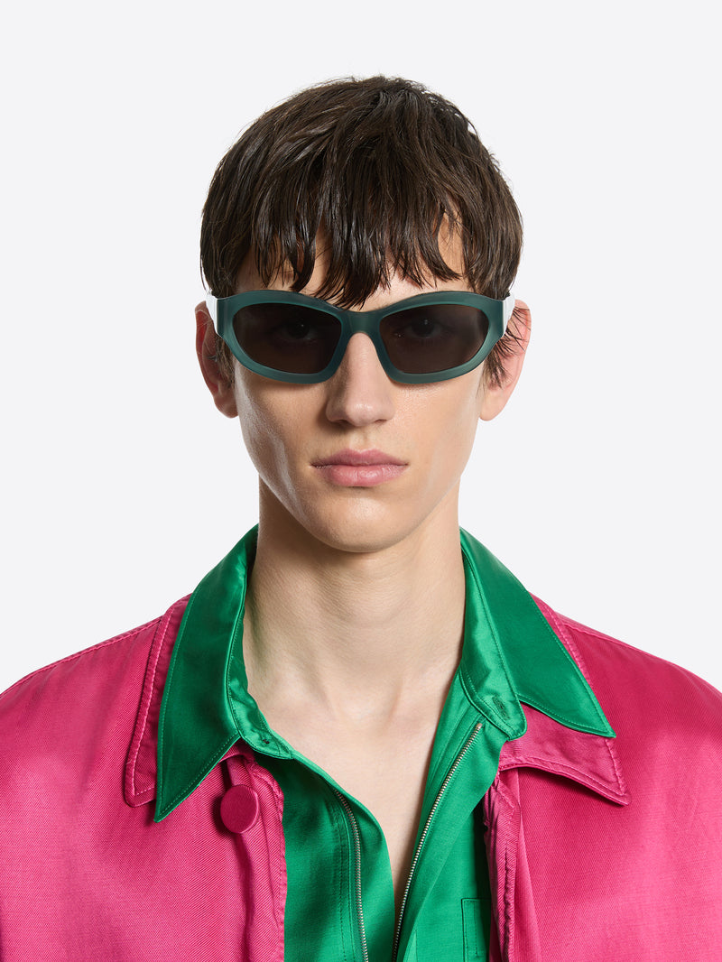 Person wearing green sunglasses and a pink jacket with a green shirt underneath on a white background