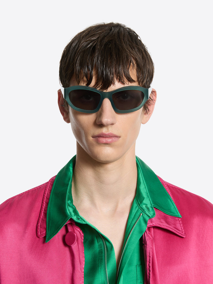 Person wearing green sunglasses and a pink jacket with a green shirt underneath on a white background