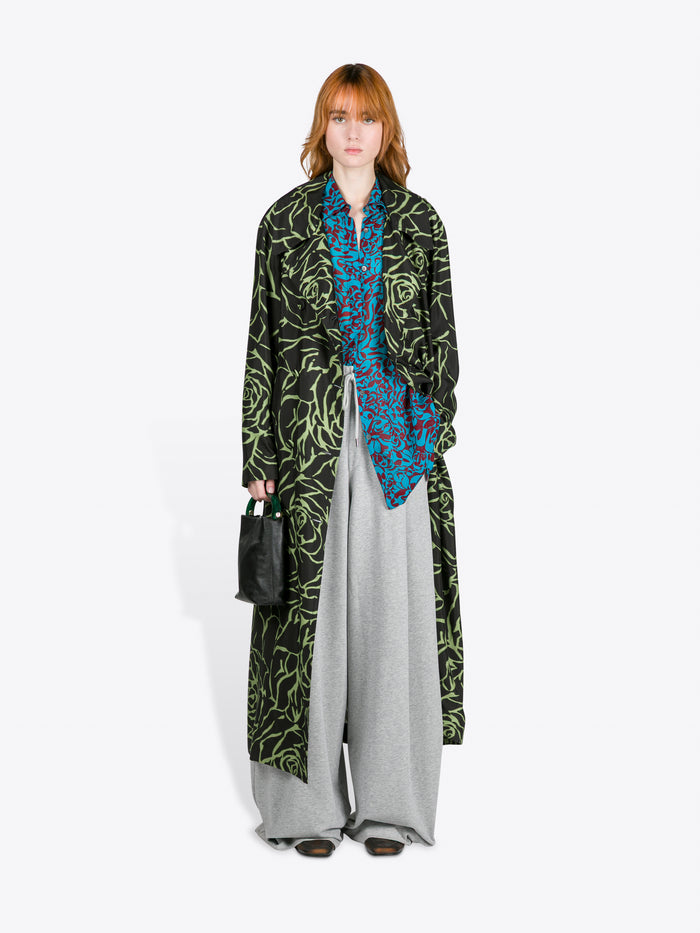 Person wearing a long black coat with green patterns, a blue patterned shirt, and gray pants on a white background. - Dries Van Noten