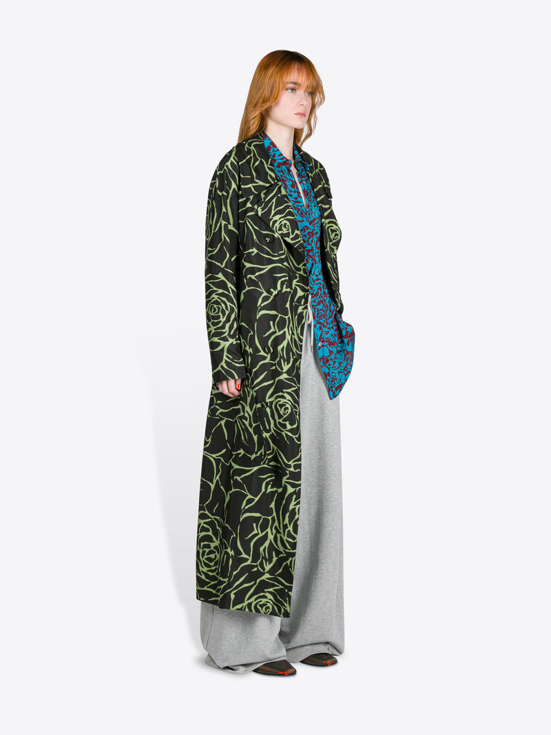 Person wearing a long, dark green patterned coat with a blue scarf on a white background - Dries Van Noten