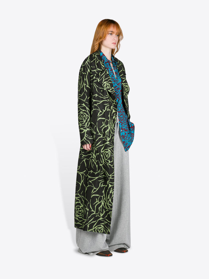 Person wearing a long, dark green patterned coat with a blue scarf on a white background - Dries Van Noten