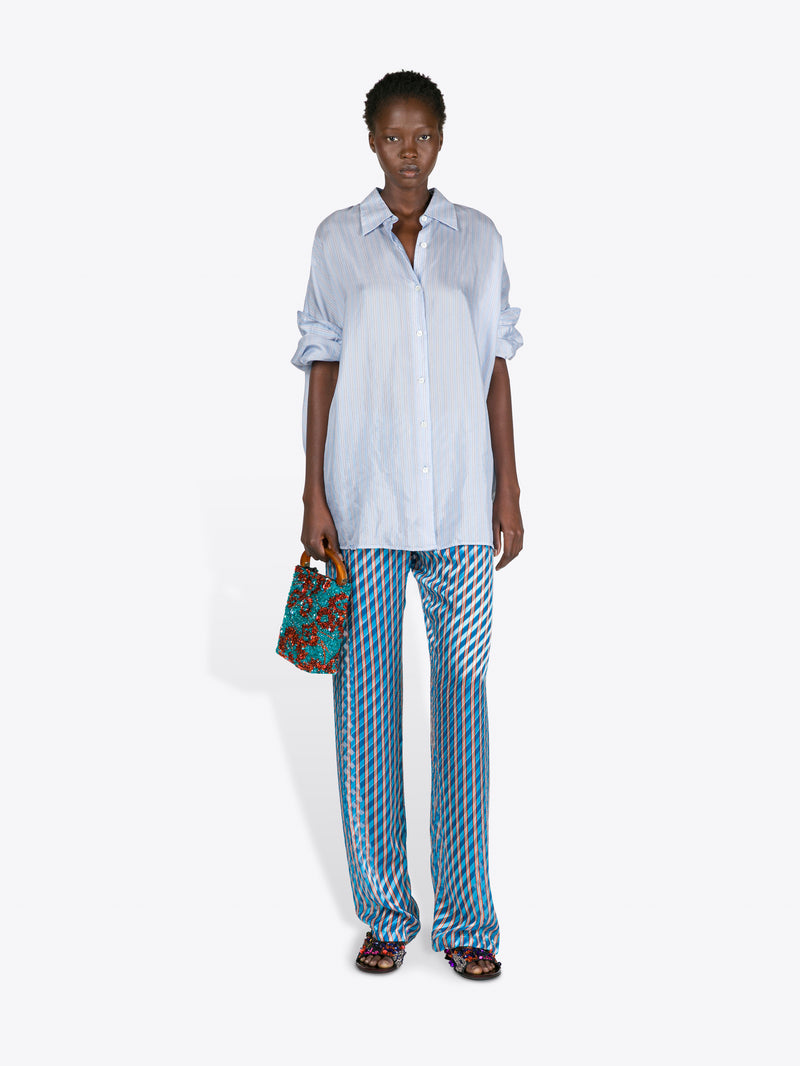 Woman wearing a light blue shirt and patterned pants, holding a colorful handbag on a white background