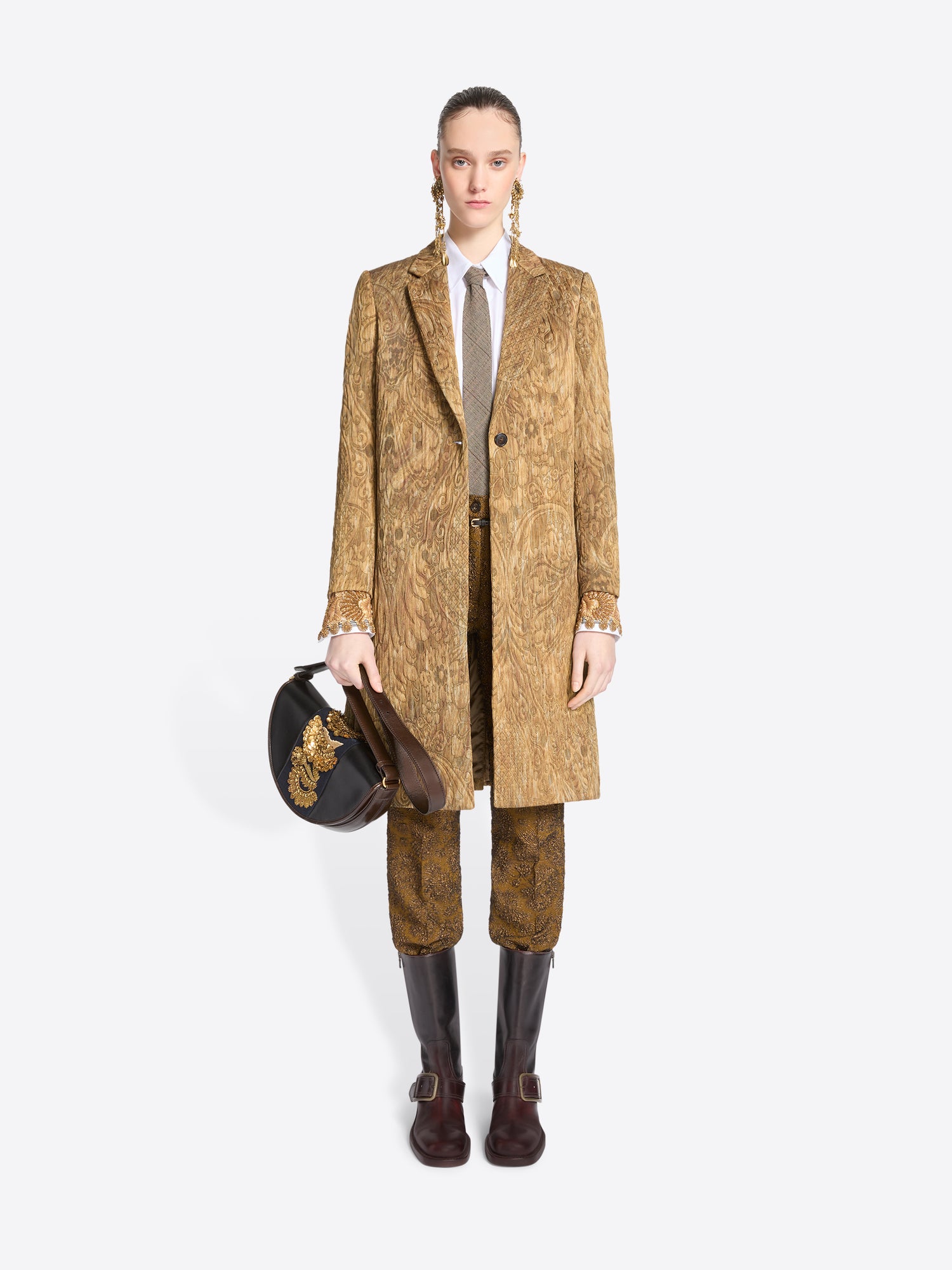 Full-length model wearing a gold paisley coat and matching trousers with a white shirt, gray tie, long gold-toned earrings, and dark brown boots, carrying a black half-moon bag with ornate gold embroidery.
