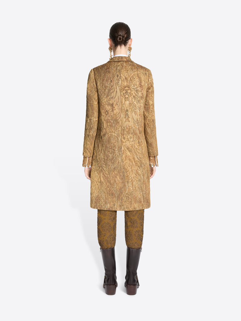Back view of a model standing against a light gray background wearing a long tan patterned coat with patterned trousers and dark brown boots.