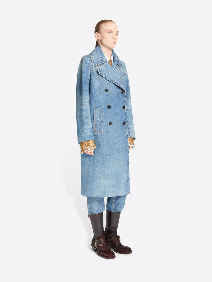 A model stands against a light gray background wearing a long light blue denim double-breasted coat with a wide, ornate patterned belt, matching light blue pants and dark brown boots.