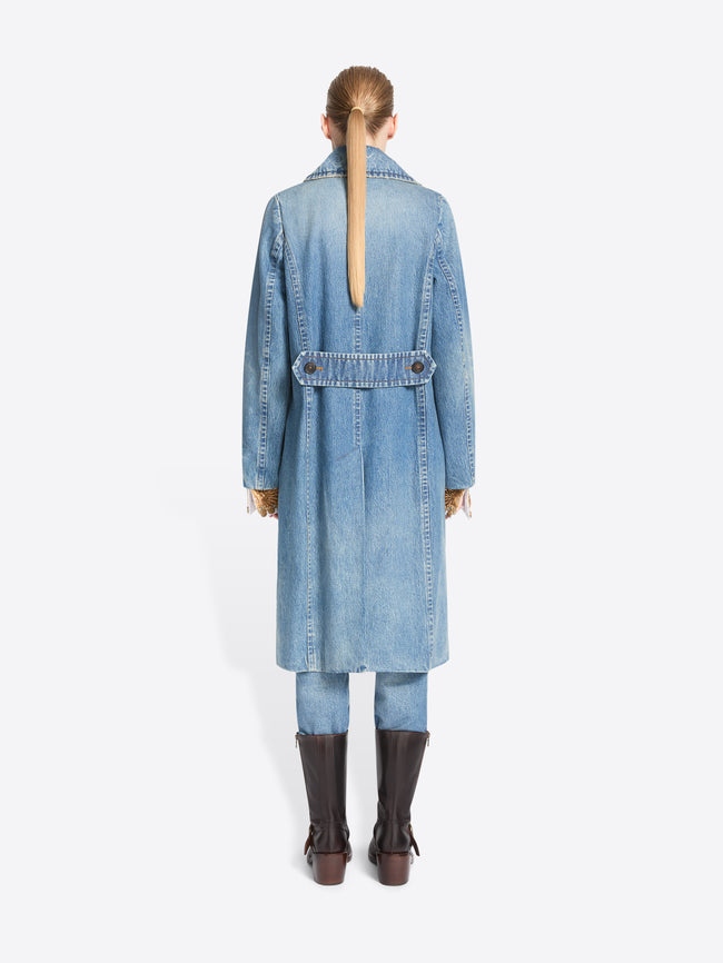 Back view of a model standing against a light gray background wearing a long light blue denim double-breasted coat with a wide, ornate patterned belt, matching light blue pants and dark brown boots.