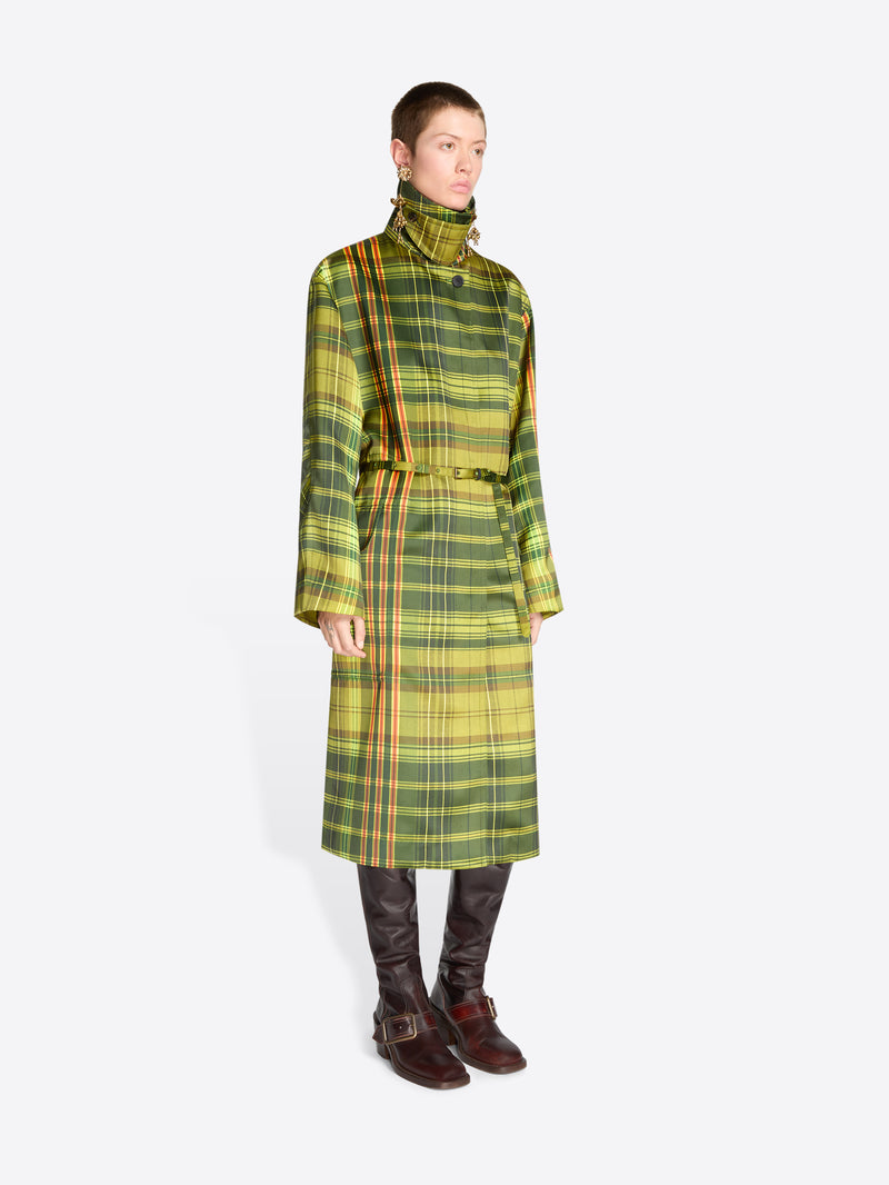 A model stands against a light gray background wearing a long green and yellow plaid coat with a high collar and waist tie and dark brown boots. 