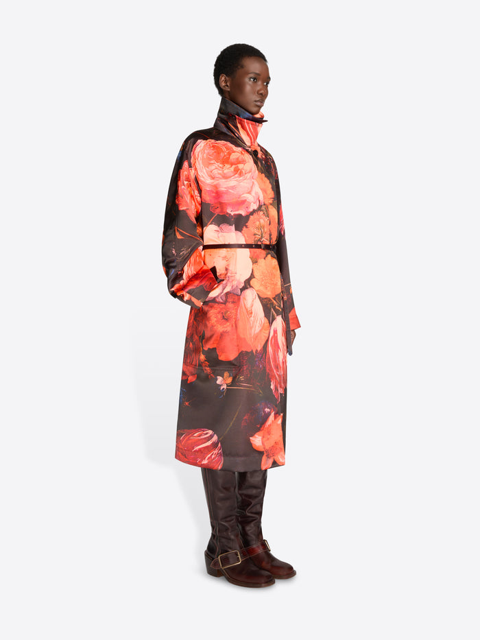 A model stands against a light gray background wearing a long-sleeved high-neck belted coat, paired with dark brown boots.