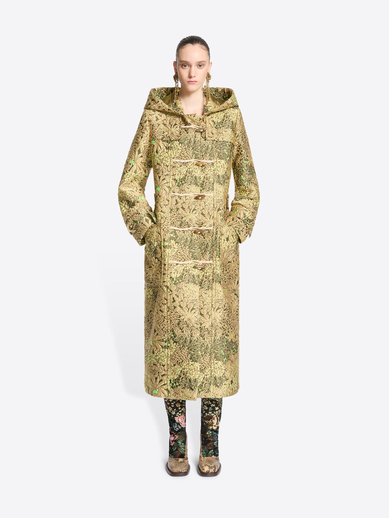 A model wearing a long hooded duffel coat with an intricate olive green and beige pattern, paired with matching patterned tights and shoes, set against a light gray background.