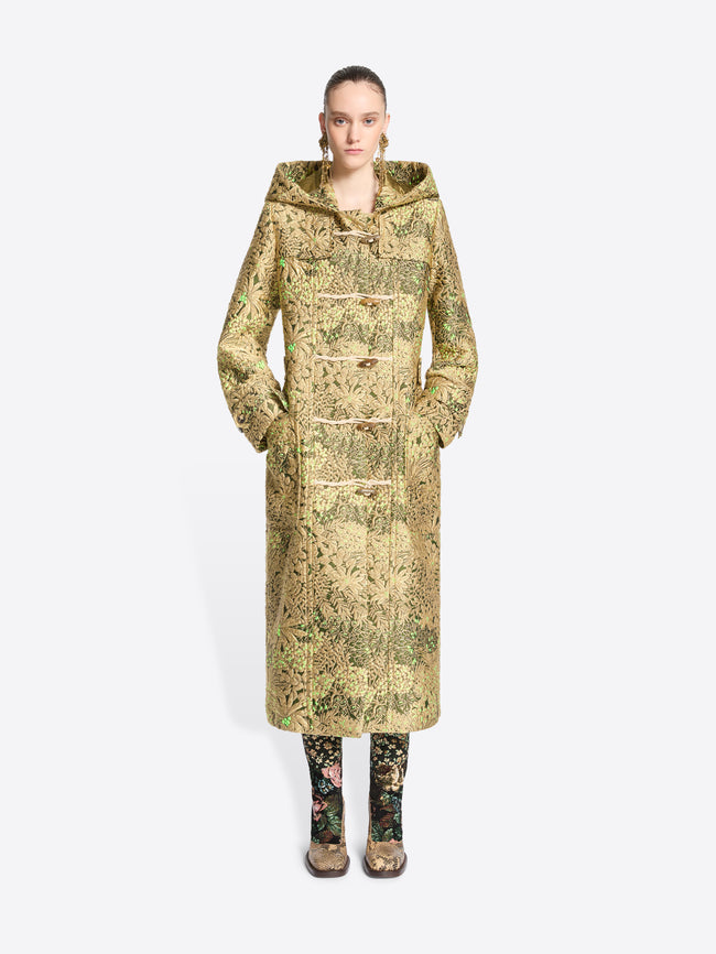 A model wearing a long hooded duffel coat with an intricate olive green and beige pattern, paired with matching patterned tights and shoes, set against a light gray background.