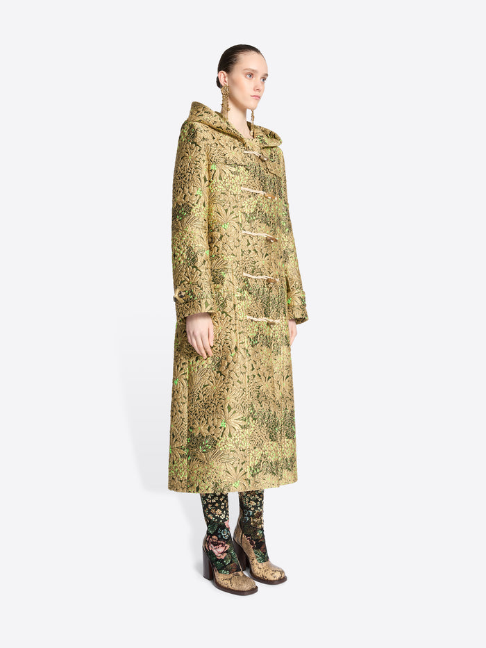 A model wearing a long hooded duffel coat with an intricate olive green and beige pattern, paired with matching patterned tights and shoes, set against a light gray background.