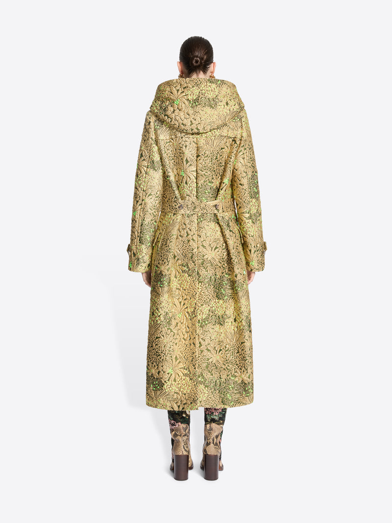 Back view of a model wearing a long hooded duffel coat with an intricate olive green and beige pattern, paired with matching patterned tights and shoes, set against a light gray background.