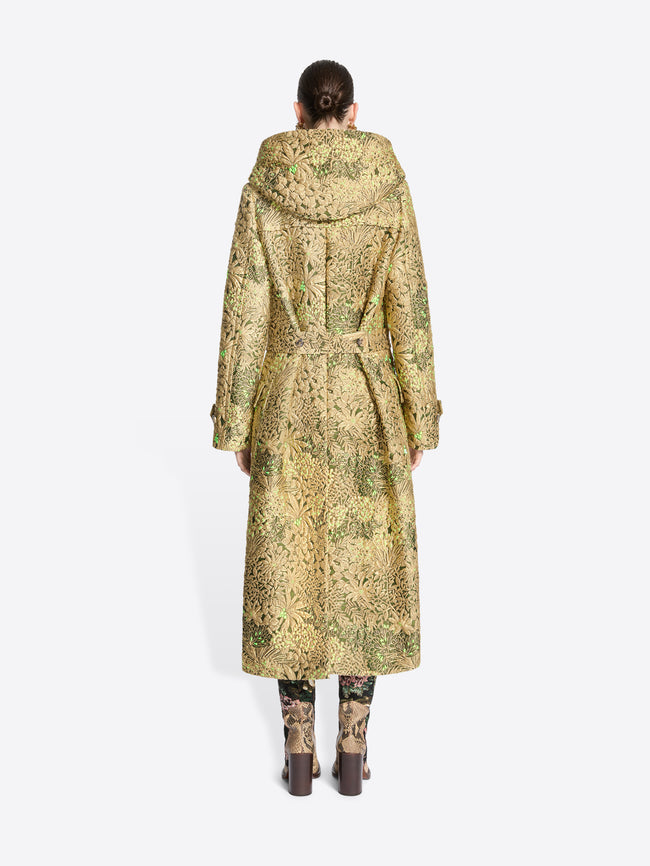 Back view of a model wearing a long hooded duffel coat with an intricate olive green and beige pattern, paired with matching patterned tights and shoes, set against a light gray background.